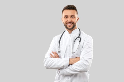 Front view of attractive confident brunette male doctor wearing lab coat and stethoscope, looking at camera with happy facial expression. Close up of intern isolated on grey. Concept of medicine.